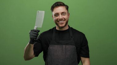 Önlük takan sakallı genç adam eldivenli bir baltayı yeşil bir duvara doğru tutuyor ve sırıtıyor..