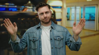 Sıradan kot ceketli sakallı genç adam spor salonunda arka planda bulanık spor ekipmanlarıyla fitness ortamını öneriyor.