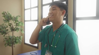 Yeşil önlüklü genç doktor gülümsüyor ve telefonda, parlak bir hastane odasında stetoskop ve kimlik kartıyla konuşuyor..