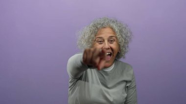 Gri saçlı yaşlı kadın gülüyor ve canlı mor bir arka plana sevinçle işaret ediyor..