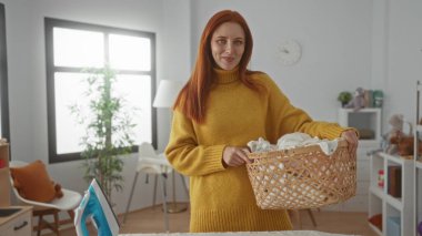 Çamaşır sepetini, parlak pencereli, kırmızı saçlı, sarı süveterli ve modern dekorlu ev işi sahnesini gösteren rahat bir odada tutan bir kadın..