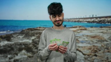 Deniz kenarında Rus rublesi tutan adam, güneşli bir günde, plajın canlı manzarası ve ışıl ışıl denizle çevrili düşünceli bir ifadeyle..