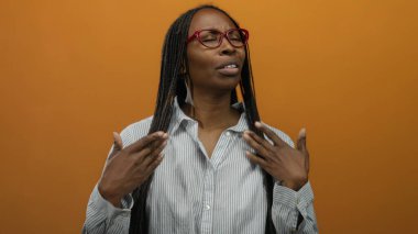 Gözlüklü ve uzun saçlı bir kadın turuncu bir duvara dayanıyor. Çizgili bir gömlek giyiyor. Rahat ve huzurlu görünüyor..