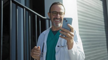 Bir şehir ortamında hastane dışında akıllı telefon kullanırken gülümseyen sağlık ceketli İspanyol kıdemli bir adam profesyonel bir bağlantı ve pozitiflik duygusunu aktarıyor..