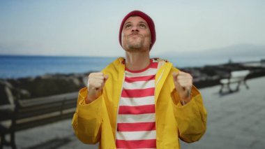 Kırmızı bereli ve sarı yağmurluklu genç adam arka planda okyanus manzaralı deniz kenarında gezinti yapmaktan zevk alıyor..