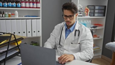 Klinikte dizüstü bilgisayarda çalışan İspanyol erkek doktor, sağlık hizmetlerinde yoğunluk ve odak gösteren tıbbi ekipmanlarla birlikte.