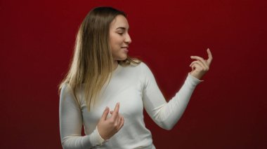 Genç sarışın kadın canlı kırmızı bir arka planda gitar çalarken şakacı bir şekilde taklit ediyor, izole bir ortamda rahat ve neşeli bir yüz ifadesi sergiliyor..