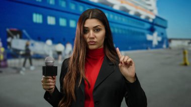 İspanyol kadın muhabir, arka planda yolcu gemisi olan bir limanda mikrofon tutuyor. Ciddi bir ifadeyle el kol hareketi yapıyor ve işaret parmağını kaldırıyor..
