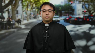 Bulanık araçları ve yayaları olan, gözlük takan ve haçlı büro kıyafetleri giyen, yoğun şehir geçmişiyle gün ışığında şehrin dışında duran bir rahip.