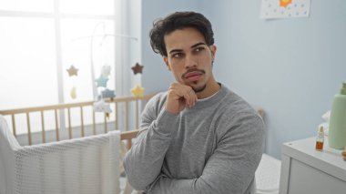 Bir bebeğin beşiğinin yanındaki yatak odasında Latin kökenli genç bir adam. Düşünceli bir yüz ifadesi ve yumuşak bir gün ışığı.
