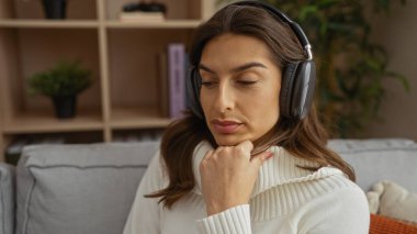 Kulaklık takan bir kadın modern bir apartman dairesinde rahat bir koltukta dinleniyor. Zarif dekorlu ve hafif ışıklandırmalı huzurlu ve rahat bir ortam yaratıyor..