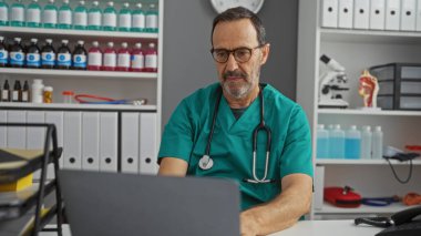 Yeşil önlüklü olgun İspanyol bir adam, profesyonel bir sağlık hizmeti için raflarla çevrili bir tıbbi klinikte dizüstü bilgisayarın başında çalışıyor.