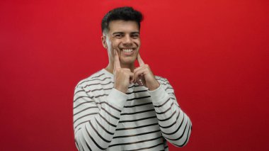 Genç adam dalgalı yanaklarıyla neşeli jestler yapıyor çizgili süveter giyiyor canlı kırmızı arka plana karşı mizahi ifadeler ve portre ortamında neşeli bir ruh hali ima ediyor..