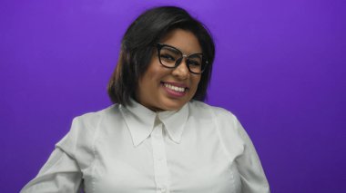 Gözlüklü genç Latin kadın sıcak bir şekilde gülümsüyor, canlı mor bir arka plana karşı jest yapıyor, şık beyaz bir gömlek giyiyor, pozitiflik ve kendine güven yayıyor..