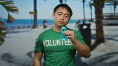 Mavi kurdeleli ve gönüllü tişörtlü genç adam güneşli bir sahil yürüyüşünde duruyor, sakin su ve yeşillikle deniz kenarında kanser farkındalığını vurguluyor..