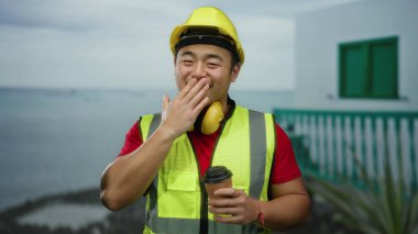 Güvenlik yeleği giyen ve kasketli bir adam arka planda bina olan deniz kenarında kahve tutuyor.
