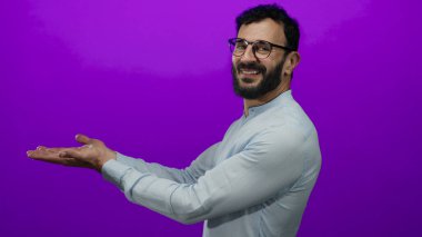 Gözlüklü İspanyol bir adam canlı mor bir arkaplan karşısında el kol hareketi yapıyor, bir sunum veya ürün sergiliyor davetkar bir gülümseme ve gündelik stil ile.