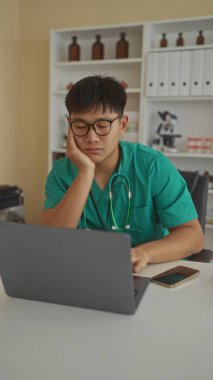 Steteskoplu yeşil önlüklü genç doktor, tıbbi ekipman ve raflarla çevrili modern bir klinikte dizüstü bilgisayar üzerinde çalışırken düşünceli görünüyor..