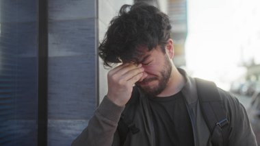 Sakallı genç adam güneşli bir günde dışarıda, yüzünü elleriyle kapatarak şehir ortamında modern bina zeminine karşı stres saçıyor..