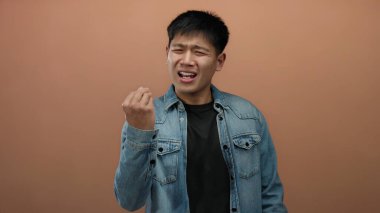 İzole edilmiş bir kahverengi duvar arkaplanına el hareketleriyle şüphecilik gösteren genç bir adam, günlük bir kot ceket giyerken belirsizlik ve tefekkür taşıyor.