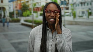 Kıvırcık saçlı, gülümseyen bir kadın şehir sokaklarında jest yapıyor.