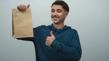 Mavi kapüşonlu ve gülümserken olumlu jestler yapan, izole edilmiş beyaz arka planda kağıt çanta tutan genç İspanyol adam..