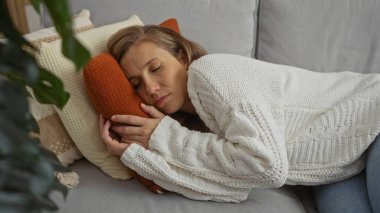 Kadın rahat bir oturma odasında, rahat bir kanepede uyuyor, beyaz kazak ve kot pantolon giyiyor, yastıklarla çevrili, sıcak, rahat bir ev ortamı öneriyor..