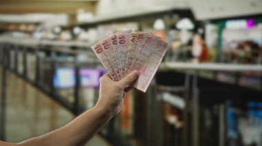 Kapalı bir alışveriş merkezinde alışveriş yaparken elinde Türk lirası banknotları tutan bir adam kalabalık bir pazar atmosferini yansıtan çeşitli dükkanlar gösteriyor..