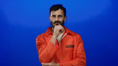 Turuncu hapishane tulumu giyen İspanyol sakallı adam mavi arka planda duruyor, eli çenesinde dalgın görünüyor..