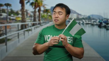 Deniz kenarındaki gezinti güvertesinde Nijerya bayrağı taşıyan genç bir adam, gerginliğini dile getiriyor, plaj kenarında açık havada gönüllü gömleği giyiyor..