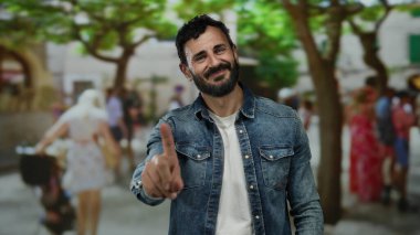 Sakallı İspanyol bir adam, arka planda bulanık insanlar ve ağaçlarla dolu kalabalık bir sokakta dikilirken 