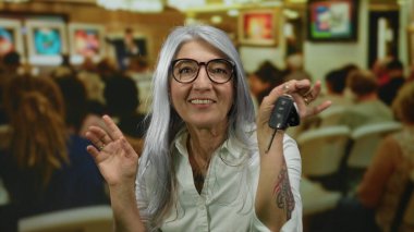 Sanat galerisinde elinde anahtarlarla gülümseyen gri saçlı yaşlı bir kadın. Etrafı insanlarla ve renkli resimlerle çevrili..