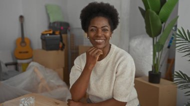 Konforlu, yeni bir oturma odasında gülümseyen bir kadın kutuları açılmış, köşede gitar, yanında büyük bir bitki, modern apartman pencerelerinden süzülen parlak bir öğleden sonra ışığı..