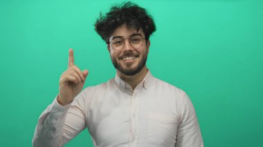 Sakallı genç adam yeşil bir arka plana karşı yukarıya doğru bakıyor, merak ve pozitiflik yayıyor..