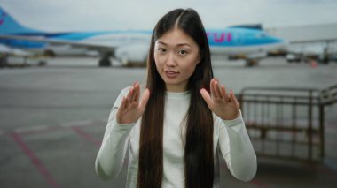 Havaalanı terminalinde sakince el kol hareketi yapan bir kadın, arka planda bir uçak, sakin bir seyahat atmosferini vurguluyor..