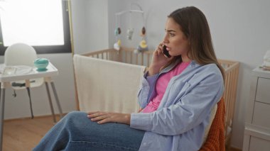 Bebek odasında rahat bir şekilde konuşan, etrafı beşik ve dekorla çevrili, evde huzurlu bir aile ortamı sergileyen bir kadın..