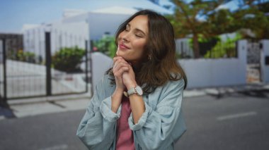 Genç İspanyol kadın güneşli bir şehir caddesinde sıcak bir şekilde gülümsüyor, neşeli kişiliğini şehir yeşilliği ve beyaz çitlere karşı sergiliyor..