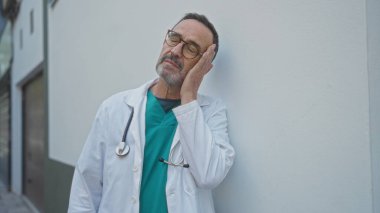 Doktor önlüklü İspanyol bir adam şehir sokağında dikiliyor. Endişeli bir ifadeyle yüzüne dokunuyor. Açık hava tıp mesleğinin baskılarını somutlaştırıyor..