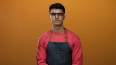 Gözlüklü ve önlüklü genç adam sağlam turuncu bir arka plana karşı duruyor, sıradan bir ortamda şık ve profesyonel bir aura yayıyor..