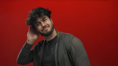 Sakallı genç İspanyol bir adam cesur bir kırmızı arka plana karşı kulak veriyor, merak ve konsantrasyon taşıyor..