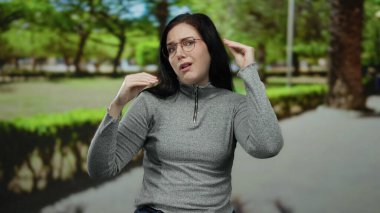 Kumral saçlı ve gözlüklü bir kadın açık havada, yemyeşil bir parkta duruyor, el hareketleriyle ve düşünceli bir bakışla kafa karışıklığını ifade ediyor, gri bir bluz giyiyor..