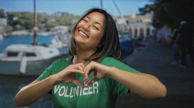 Genç bir kadın arka planda teknelerle limanda kalp hareketi yapıyor yeşil gönüllü tişörtü giyerken gülümsüyor ve açık deniz manzarasını yakalıyor..
