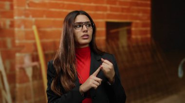 İç mekan inşaatında gözlüklü genç İspanyol kadın profesyonel siyah ceket giymiş kırmızı tuğla bir duvarın önünde tutkuyla konuşuyor..