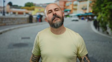Sakallı ve dövmeli kel adam şehir sokağında duruyor. Arka planda bulanık binalar var. Yakışıklı ve açık hava havasını yansıtıyor..