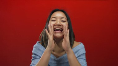 Kadın canlı kırmızı duvar arka planına karşı bağırıyor. Yüzüne yakın ellerle heyecanını dile getiriyor. Mavi süveter giyiyor, Çin kültüründen soyutlanmış..