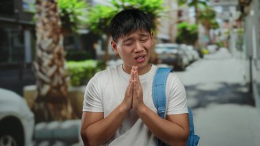 Sırt çantalı genç bir adam güneşli bir şehir caddesinde durup dua ederken palmiye ağaçları ve park halindeki arabaların arasında duygularını ifade ediyor..