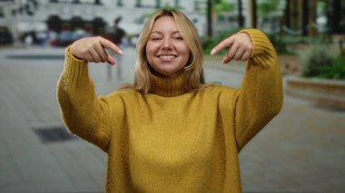 Sarı süveterli bir kadın şehir sokaklarını işaret ediyor güneşli bir günde şehrin arka planında neşeli hareketler sergiliyor..