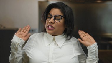 İçerde gözlüklü ve beyaz gömlekli bir kadın, elleri hafif havada, ağzı kapalı ve gözleri kapalı, sakin bir tavır sergileyen bir ifade sergiliyor.