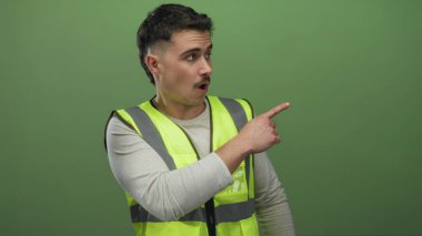 Yansıtıcı yelek giyen genç İspanyol adam, izole edilmiş yeşil duvar arka planına karşı şaşırmış bir ifade sergiliyor..