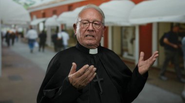 Siyah cüppeli yaşlı rahip, arka planda insanlarla dolu kalabalık bir sokakta tutkuyla el kol hareketi yapıyor..